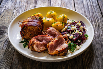 Medium well done pork loin wrapped in bacon with fried potatoes and feta cheese on beets on wooden table
