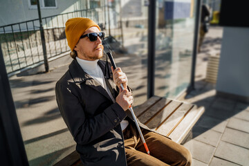 Blind man with a walking stick sitting on a bench at a public transport stop