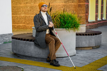 Blind man with a walking stick sitting on a bench in the city