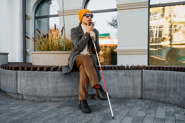 Blind man with a walking stick sits on a bench uses a smartphone
