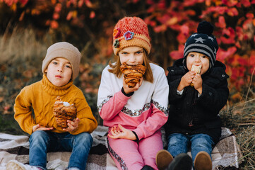 A fashionable preschool boy, a boy child and his friend a smiling girl eat a croissant, a bun, sitting on a plaid in autumn in nature. Family, friendship, brother and sister, photography.