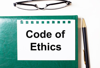 On a light background, glasses, a pen, a green notebook and a white sheet of paper with the text CODE OF ETHICS