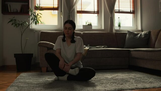Meditation at home. Young pretty Caucasian woman wearing sports clothes sits cross-legged on floor in living room. Sofa and windows at background. Concept of yoga, harmony and balance.