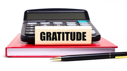 A burgundy notebook on a white background. On it is a black calculator, a pen and a wooden block with a GRATITUDE text. Business concept