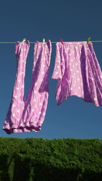 Vertical video social media format – Closeup of the two parts of a wet pair of pink with white spots pyjamas / pajamas, hanging by pegs and blowing dry on a garden / yard washing line.