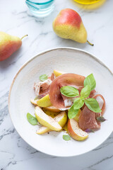 Prosciutto ham with pear wedges and fresh green basil in a beige plate, vertical shot on a white marble background