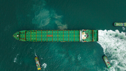 tugboat dragging cargo ship leaving shipyard after repairs in green sea, aerial top view