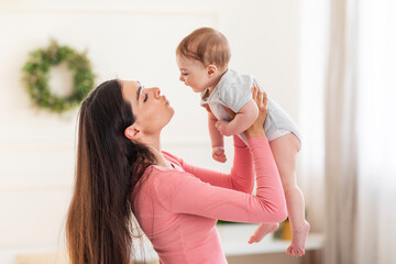 Young mom lifting her adorable newborn baby in air while standing in bedroom at home, holding him...