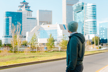 Side view of tourist viewing, enjoying modern architecture of skyscrapers. Young man wearing jacket and hat standing on street against backdrop of city