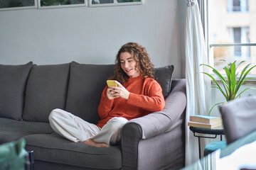 Young woman sitting on couch holding smartphone, looking at cellphone using cell phone checking...