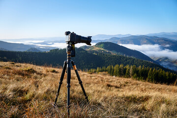 Camera on tripod on the meadow in the mountains capturing a beautiful mountain landscape with a fog, clouds and a forest. Landscape photography