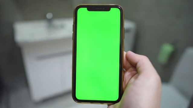 Use green screen for copy space closeup. indoors of toilet or bathroom. Chroma key mock-up on smartphone in hand. young man holds mobile phone iPhone and swipes photos or pictures