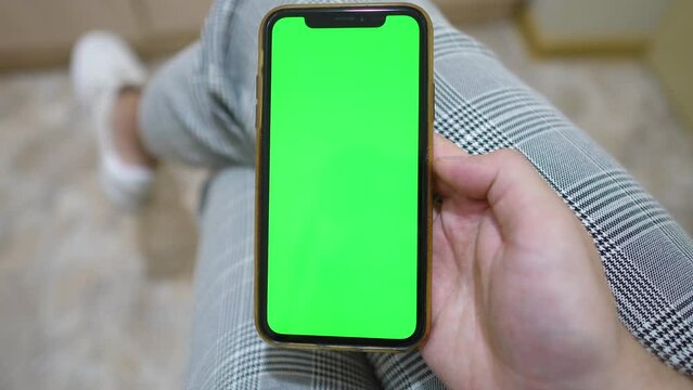 Use green screen for copy space closeup. in front of a desktop with a laptop. Chroma key mock-up on smartphone in hand.  young man holds mobile phone iPhone and swipes photos or pictures