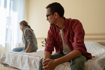 Young unhappy family couple not talking after quarrel, sitting separately on bed discussing breakup divorce. Frustrated man husband looking at crying wife sitting aside. Problems in bad relationship