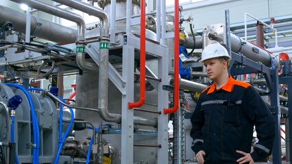 A young inexperienced engineer at his workplace in a white helmet and overalls inspects the equipment of the compressor station. Work at an industrial plant in an industrial workshop.