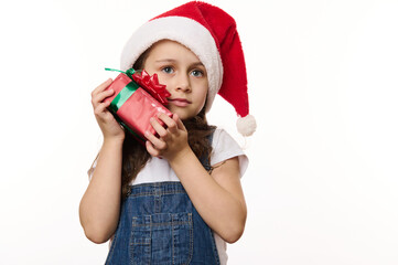 Close-up portrait of charming little child, cute baby girl wearing Santa hat, with a Christmas present, isolated on white background with copy ad space. Looking forward to the upcoming winter holidays