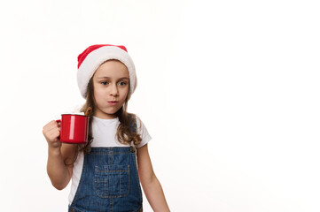 Mischievous lovely baby girl wearing Santa hat and denim overalls, drinking hot cocoa drink and eating sweet marshmallows, isolated on white background with copy ad space. Magic Christmas atmosphere
