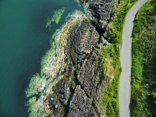 Fototapeta premium Aerial view of a coastal path with rocky cliffs and lush greenery by the calm sea in Bangor, Ireland
