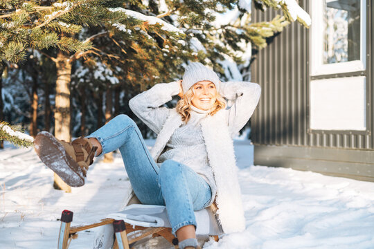 Happy woman with blonde hair in winter clothes having fun with sleigh against background of snowy winter forest