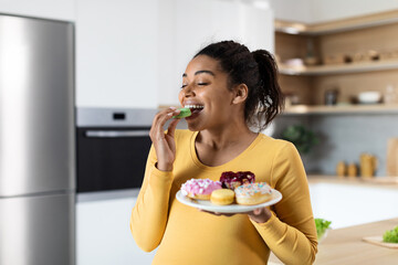 Smiling hungry young black pregnant female with big belly eats sweets, hold plate with cookies - obrazy, fototapety, plakaty