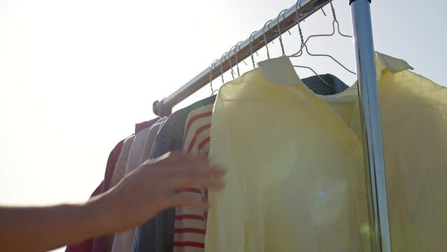 Woman hand choosing pre owned clothes hanging on rail, close up view, outdoors against sky.