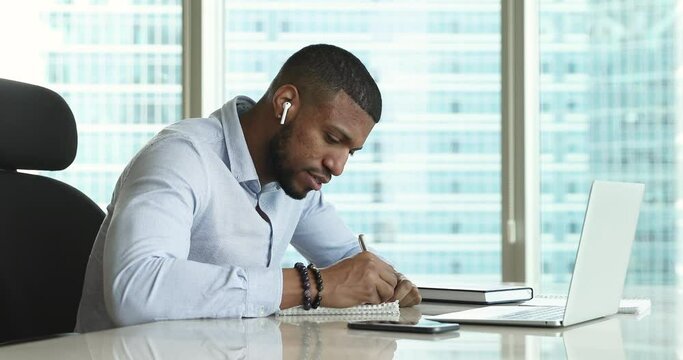 Focused African student, intern guy with wireless earphone in ear writing notes at laptop, watching learning webinar, talking to teacher on video call. Employee giving online customer support
