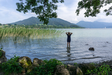 Fototapeta na wymiar Eine junge Frau steht am Ufer des Tegernsees im Wasser und schaut hinaus auf die Berge