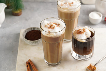 Three tall glasses with warm coffee drink with cinnamon, whipped milk foam and caramel on a marble board and christmas ornaments and decoration