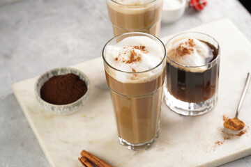 Three tall glasses with warm coffee drink with cinnamon, whipped milk foam and caramel on a marble board and christmas ornaments and decoration