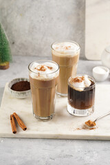 Three tall glasses with warm coffee drink with cinnamon, whipped milk foam and caramel on a marble board and christmas ornaments and decoration