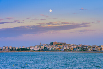 Vieste, Italy. View of the town of Vieste after sunset at dusk with the moon high in the sky. Above, the Swabian Aragonese Castle with the historic center below. September 9, 2022.