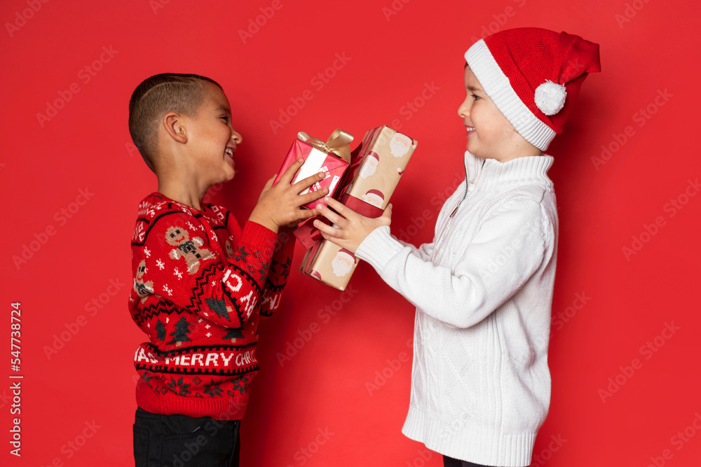 Wall mural Two cute friends have fun with christmas gifts in studio on red background.