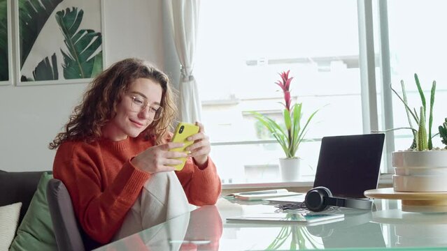 Young happy smiling pretty woman sitting on chair holding smartphone using cell phone technology, looking at mobile phone sitting at home table, texting messages, doing mobile shopping, buying online.