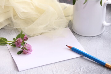 Empty note paper sheet with pencil. A sprigs of clover on the table and on the mug, mesh fabric texture cloth. Sweet moments in cozy home.
