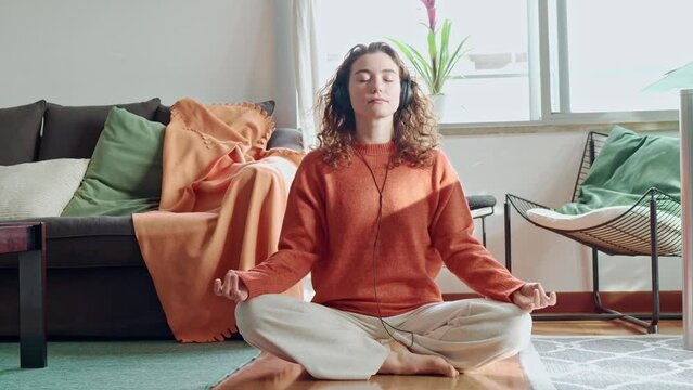 Young pretty healthy mindful woman wearing headphones listening calming music audio podcast sitting on floor at home doing yoga meditation breathing exercise for mental balance feeling peace of mind.
