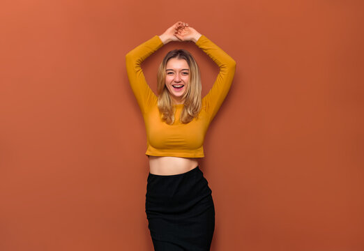 A beautiful young girl with large breasts, holds her hands above her head, laughs at the camera with her eyes narrowed, in a yellow blouse with bare belly and a black long skirt on an orange backdrop.