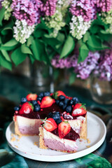 Cheesecake with strawberries and blueberries, close-up, in a rakoze, against a background of lilacs. Vertical orientation.