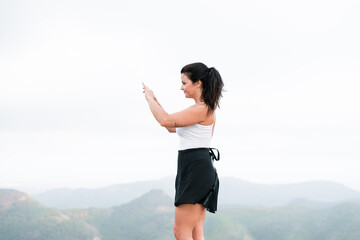 brunette caucasian mature woman in white top and black skirt making a photo of herself with her smartphone on top of mountain