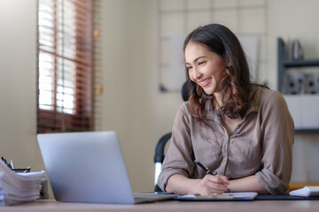 Young Asian business woman sitting happily working on a laptop take notes carefully and smile happily on assignment at home.