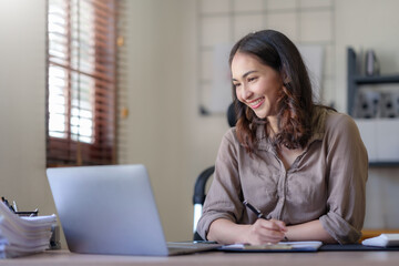 Obraz premium Young Asian business woman sitting happily working on a laptop take notes carefully and smile happily on assignment at home.