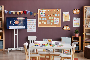 Interior of spacious classroom for nursery schoolkids with table surrounded by chairs in center and boards with notes and pictures