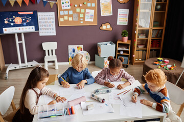 Group of creative little learners of nursery school gathered by table drawing with multi-color...