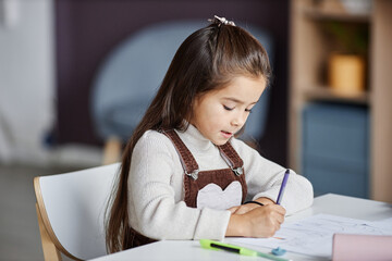 Diligent little girl with dark long hair drawing with crayons on paper while sitting by table in...