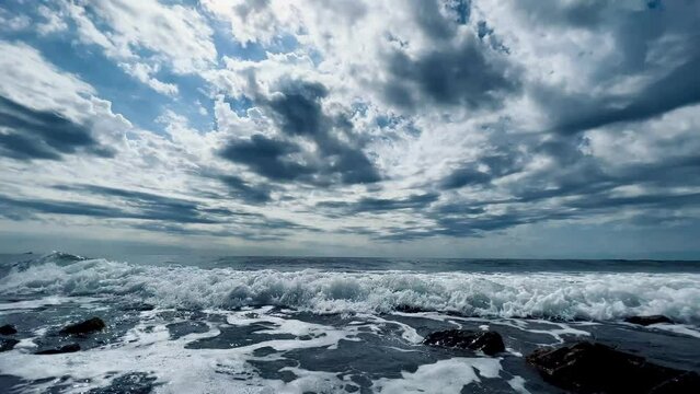 Daytime panorama of the camera from top to bottom from the cloudy sky to blue clean sea. Active fast waves roll onto shore, water with white sea foam seeps among stones. Close to water. 4k footage