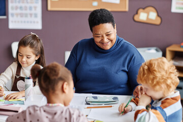 Focus on happy African American mature teacher and little learner with dark long hair sitting in front of two intercultural nursery schoolkids
