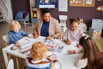 Group of intercultural happy little learners and African American mature teacher interacting at...