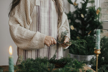 Making Christmas rustic wreath. Woman hands holding cedar branches and making wreath on rustic wooden table with ribbon, golden bells, candles. Moody holiday image. Winter holiday workshop
