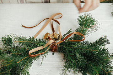 Making Christmas modern wreath. Woman hands decorating wreath with golden bells and ribbon on white wooden table. Holiday workshop. Winter decor, moody image