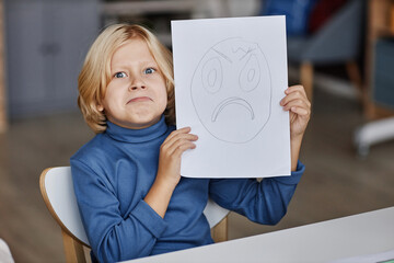 Cute blond boy imitating expression of gloomy face drawn on paper that he showing to you while...