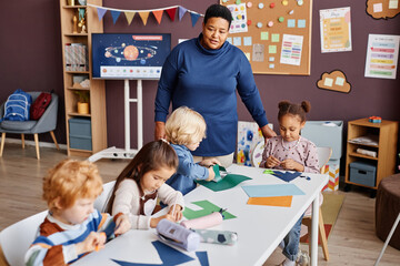 Confident teacher standing in front of table where group of little learners of primary school creating paper toys or origami at lesson